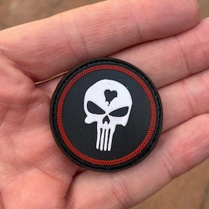 May include: A black and white embroidered patch with a white skull and crossbones with a red bullet hole in the forehead. The patch is surrounded by a red stitched border.