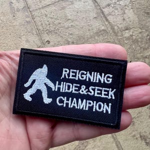 Può includere: Toppa ricamata nera con una silhouette bianca di Bigfoot e il testo "Reigning Hide & Seek Champion".