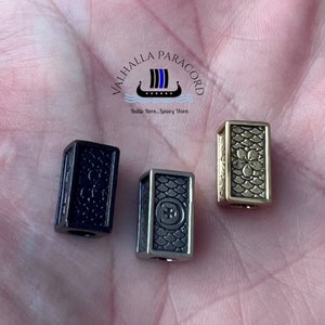 May include: Three metal paracord tips in different finishes: black, gunmetal, and gold. Each tip features a textured design with a raised logo.