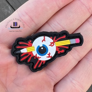 Puede incluir: Un dije de goma negra con un ojo blanco con iris azul, un lápiz amarillo y salpicaduras de sangre roja. El dije tiene el texto "Valhalla Paracord" y "Battle Born...luxury Worn".