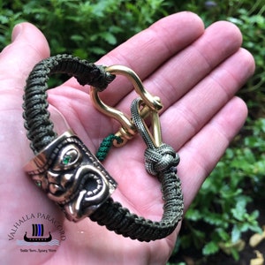 May include: Olive green paracord bracelet with a brass clasp and a decorative bronze bead featuring a face design with green accents. The bracelet is handmade and features a secure knot closure.