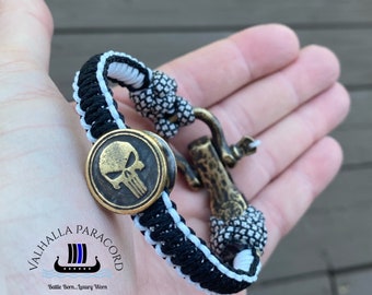 May include: Black and white paracord bracelet with a bronze-colored skull emblem and clasp. The bracelet features a braided design with a white center and black edges. The clasp is a bronze-colored hook. The text "VALHALLA PARACORD" is visible.