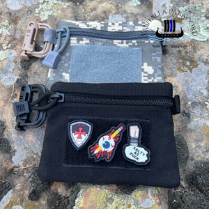 Peut inclure: Deux petites pochettes zippées avec mousquetons. L'une est noire avec trois écussons, l'autre est camouflage. La pochette noire comporte des écussons avec un emblème, un œil avec un crayon et un salière avec du texte.