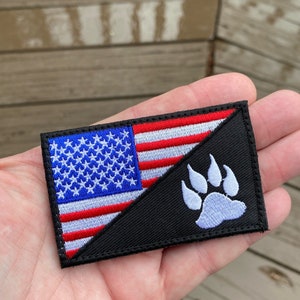 Puede incluir: Un parche bordado en blanco y negro con una división diagonal. La mitad superior presenta la bandera estadounidense con estrellas y rayas rojas, blancas y azules. La mitad inferior es negra con una huella de pata de oso blanca.