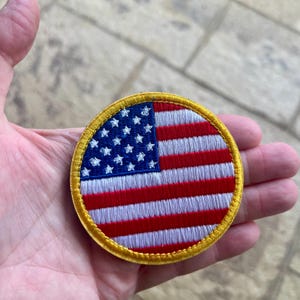 Op de afbeelding: Een ronde geborduurde patch met een gouden rand met de Amerikaanse vlag. De vlag is rood, wit en blauw met witte sterren op een blauw veld.