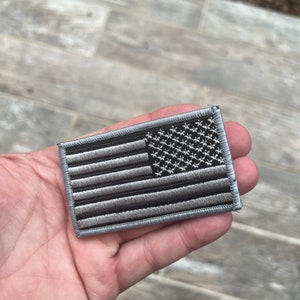 Op de afbeelding: Een rechthoekige geborduurde patch met de Amerikaanse vlag in grijstinten en zwart. Het vlagontwerp omvat sterren in de linkerbovenhoek en strepen over de rest van de patch. De patch wordt in een hand gehouden.