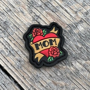 Puede incluir: Un parche bordado en negro y rojo con un corazón y una pancarta que dice "MOM". El corazón es rojo y la pancarta es dorada. Hay dos rosas rojas a cada lado del corazón.