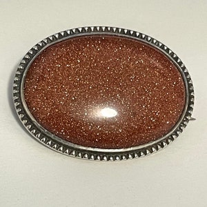 May include: A silver oval brooch with a sparkly, reddish-brown stone set in the center.