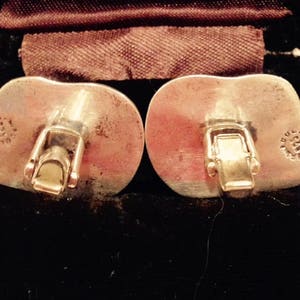 May include: A pair of silver cufflinks with a heart-shaped design. The cufflinks have a clasp closure and are marked with "925" and "MEXICO".