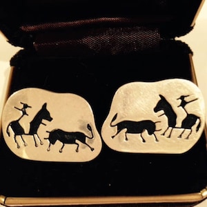 May include: A pair of silver cufflinks with a black silhouette design of a bull and horses. The cufflinks are in a black velvet box.