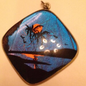 May include: A pendant with a tropical sunset design. The pendant features a blue background with an orange sun, palm tree silhouette, and a black shoreline. The pendant has a silver-colored metal frame and a small loop at the top.