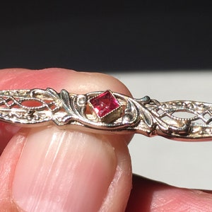 May include: A silver filigree brooch with a red square-shaped gemstone in the center.