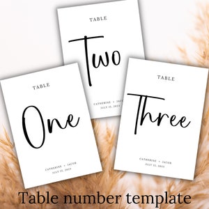 May include: Three white table number cards with black calligraphy. The numbers are "One", "Two", and "Three". Each card also includes the text "TABLE" and the names "Catherine & Jacob" and the date "July 15, 2023".