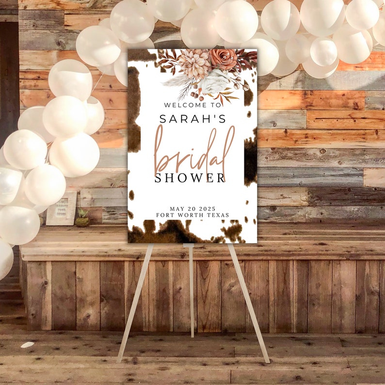 Puede incluir: Un cartel blanco con un patr&oacute;n de piel de vaca marr&oacute;n y acentos florales. El cartel dice "Bienvenido al Bridal Shower de Sarah, 20 de mayo de 2025, Fort Worth, Texas".