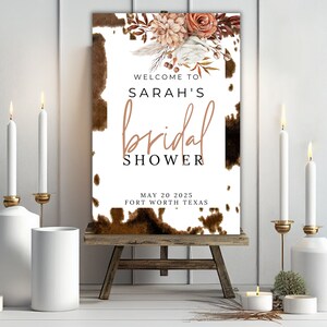 Puede incluir: Un cartel de despedida de soltera blanco con estampado de piel de vaca marr&oacute;n y detalles florales. El texto dice "Bienvenido al shower nupcial de Sarah, 20 de mayo de 2025, Fort Worth, Texas".
