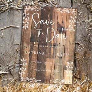 K&ouml;nnte beinhalten: Rustikales Holzschild mit dem Schriftzug "Save the Date" in wei&szlig;er Schrift. Es enth&auml;lt die Namen "Catrina + Travis" und das Datum "Samstag, 20. Mai 2025". Wei&szlig;e florale Elemente umrahmen die R&auml;nder. Der Hintergrund ist aus Stroh.