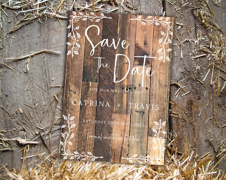 K&ouml;nnte beinhalten: Rustikales Holzschild mit dem Schriftzug "Save the Date" in wei&szlig;er Schrift. Es enth&auml;lt die Namen "Catrina + Travis" und das Datum "Samstag, 20. Mai 2025". Wei&szlig;e florale Elemente umrahmen die R&auml;nder. Der Hintergrund ist aus Stroh.