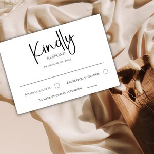 以下が含まれることがあります： 「Kindly Respond by August 30, 2023」と黒文字で書かれた白いRSVPカード。 「Joyfully Accepts」と「Regretfully Declines」の2つのチェックボックスがあります。