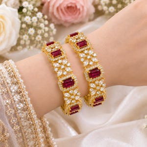 May include: Two gold-toned bangles featuring rectangular deep red stones alternating with clusters of clear marquise-cut stones. The metal settings are encrusted with small clear pavé stones, creating a detailed and ornate appearance on each bracelet.