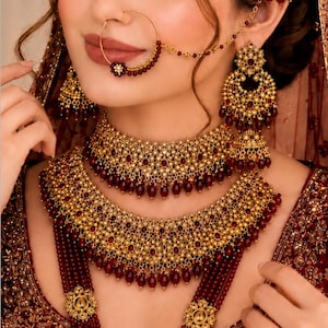 May include: A gold jewellery set featuring a multi-layered necklace, large drop earrings, a maang tikka, and a nose ring with a chain. The pieces are crafted with intricate gold metalwork, small white pearls, and deep red beads in a matching design.