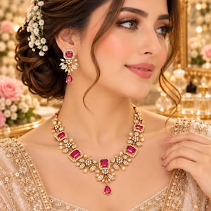 Op de afbeelding: Een goudkleurige ketting en bijpassende oorbellen met rechthoekige fuchsia-roze edelstenen, kleine witte parels en heldere pavé-kristallen in een bloemmotief, afgewerkt met een centrale druppelvormige roze steen in het midden.