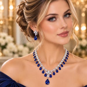 Puede incluir: Un conjunto de joyería compuesto por un collar y pendientes a juego. El collar presenta una serie de piedras azules en forma de lágrima rodeadas de cristales transparentes, con un colgante central. Los pendientes reflejan el diseño de lágrima.