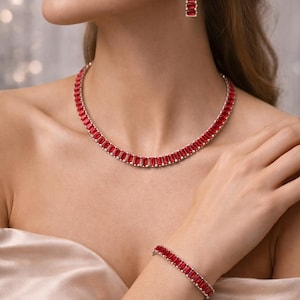 May include: A jewellery set comprising a necklace, bracelet, and earrings. Each item is embellished with rectangular, vivid red gemstones set in a silver-toned metal. The necklace encircles the neck, the bracelet is on the wrist, and the earrings hang down.