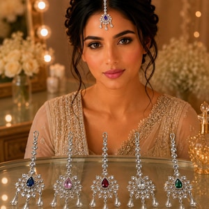 Peut inclure: Cinq bijoux de tête maang tikka argentés disposés en rangée, chacun doté d'une pierre précieuse centrale en forme de goutte bleue, rose, rouge, transparente ou verte, entourée de cristaux clairs et de pampilles sur une base circulaire.