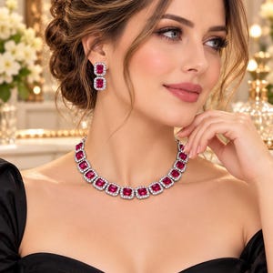 Può includere: Un set di gioielli composto da una collana e orecchini pendenti, ciascuno caratterizzato da gemme rettangolari di un vivido colore rosa-rosso, circondate da un alone di piccole pietre chiare scintillanti su una montatura argentata.