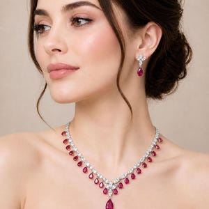 Puede incluir: Un deslumbrante conjunto de collar y pendientes con metal plateado y gemas rojas vibrantes en forma de lágrima. El collar cae con gracia, complementado por pendientes a juego, creando una estética lujosa y elegante.