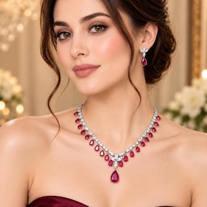 May include: A silver-toned necklace and matching drop earrings set featuring pear-shaped deep red gemstones and clear white stones. The necklace has a central teardrop pendant, while the earrings feature a floral-inspired stud design.