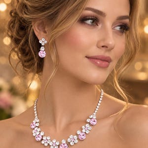 May include: A jewelry set consisting of a necklace and matching drop earrings. The necklace features a floral-inspired design with clear white crystals and round light pink crystals. The earrings feature a matching floral motif with a pink crystal.