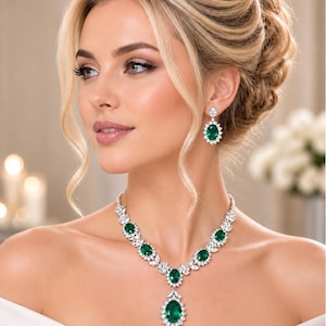 May include: A jewellery set consisting of a necklace and matching drop earrings. The necklace features oval-cut emerald green stones surrounded by clear white crystals in a silver-toned setting. The earrings mirror this design with a single oval emerald.