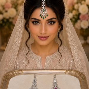 May include: A silver-toned maang tikka headpiece featuring clear crystals and green teardrop accents, paired with two matching long earrings on a tray. One earring features clear crystals, while the other features clear crystals and pink teardrop stones.