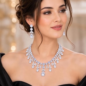 Può includere: Una collana e orecchini pendenti abbinati color argento con pietre trasparenti sfaccettate, a forma di goccia e rotonde, disposte in un motivo a cascata simile a foglie. Il set di gioielli è indossato su un capo nero con scollo ampio.