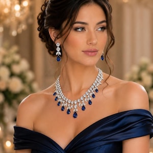 May include: A jewellery set consisting of a necklace and matching drop earrings. The necklace features a silver-toned metal base adorned with clear marquise-cut crystals and deep blue pear-shaped stones. The earrings mirror this design.