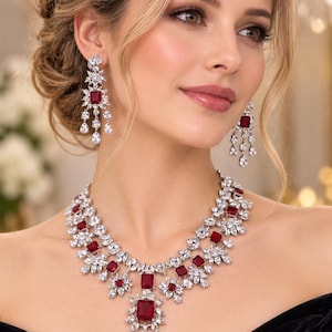 Puede incluir: Un conjunto de joyería compuesto por un collar y pendientes colgantes a juego. Las piezas presentan piedras rectangulares de color rojo intenso rodeadas de cristales transparentes en forma de marquesa y pera sobre una montura de metal plateado.