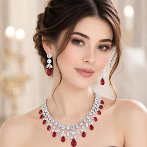 May include: A jewellery set consisting of a necklace and matching drop earrings. The necklace features a silver-toned metal base adorned with clear marquise and round-cut crystals, accented by vibrant red pear-shaped stones in a tiered design.