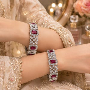 May include: Two silver-toned bracelets featuring rectangular red gemstones surrounded by small, clear, sparkling stones arranged in a floral pattern. The bracelets are wide and have a repeating design of red stones set within intricate metalwork.