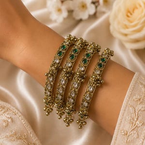 May include: A set of four gold-toned metal bangles featuring intricate filigree patterns, inlaid with teardrop-shaped green stones and clear white crystals, accented with small dangling metal beads along the edges of each circular bracelet.