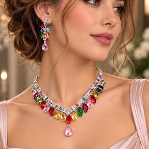 May include: A silver-toned necklace and matching drop earring set featuring a vibrant array of multi-colored, marquise-cut crystals arranged in a V-shape pattern, accented with clear baguette crystals and a single pink pear-shaped crystal pendant.