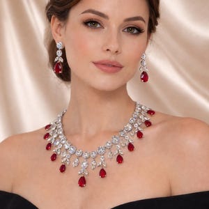 May include: A woman wearing a necklace and earrings with clear stones and red, teardrop-shaped gems. The jewellery is set against a black off-the-shoulder dress. The woman has her hair up.
