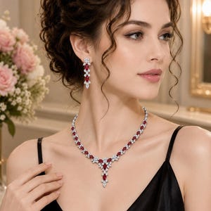 May include: A necklace and earring set with red and clear gemstones. The necklace and earrings are made of silver-coloured metal and feature oval-shaped, deep red stones, interspersed with smaller, clear stones. The jewellery is worn with a black dress.