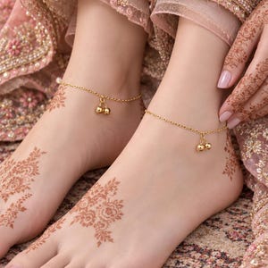 May include: A pair of gold-toned metal anklets featuring a delicate chain link design with two small, round, polished gold-toned charms dangling from the centre of each piece. The anklets are worn around the ankles, which are adorned with intricate brown henna patterns.
