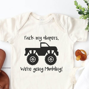 May include: A white baby onesie with a black monster truck graphic and the text "Pack my diapers, we're going mudding!"  The onesie is on a white wooden surface with a white baby blanket and a wooden rattle.