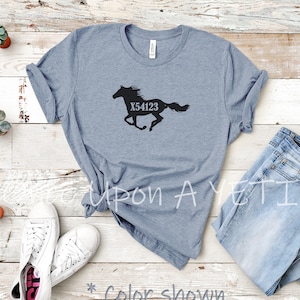 May include: Heather slate blue t-shirt with a black silhouette of a running horse and the number X54123. The shirt is displayed with a pair of white sneakers and blue jeans on a wooden surface. The text "* Color shown is Heather Slate" is also visible.