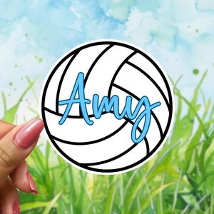 May include: A white and black volleyball sticker with the name "Amy" in light blue script. The sticker is held by a hand with pink nail polish against a watercolor background of grass and sky.