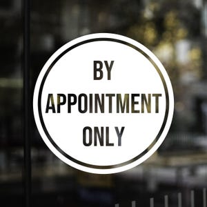 May include: A round white sign with black text that reads "BY APPOINTMENT ONLY". The sign has a black border and is likely affixed to a glass door or window. The background is blurred.