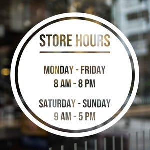May include: A circular sign with the words "STORE HOURS" at the top. Below, the sign indicates store hours: Monday-Friday 8 AM-8 PM, Saturday-Sunday 9 AM-5 PM. The sign is white with black and gold text.