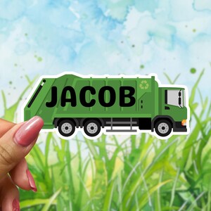 May include: A green sticker of a garbage truck with the name "JACOB" in black block letters. The truck has a recycling symbol and is set against a watercolor background of grass and sky.